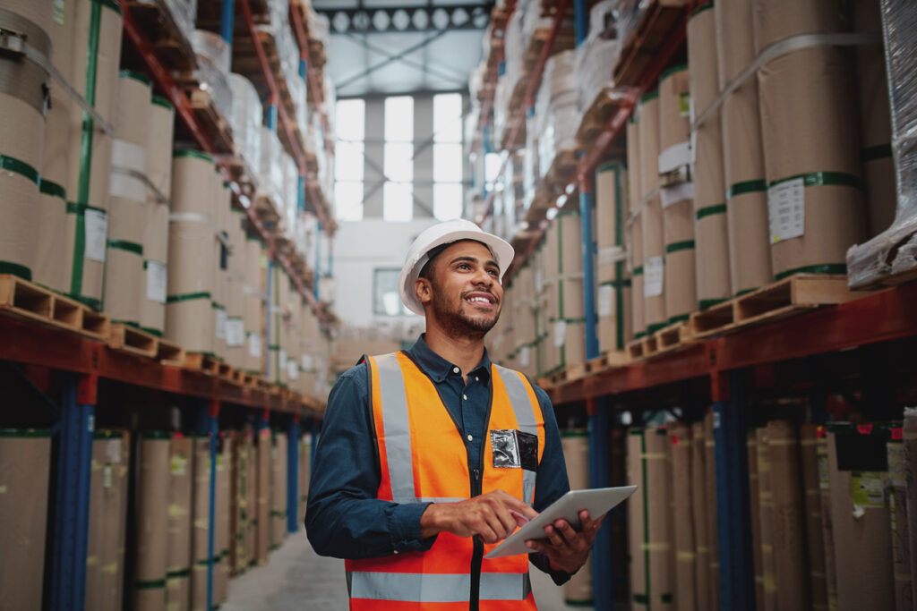 Tangentia | Low angle view of young african man wearing reflective jacket holding digital tablet standing in factory warehouse smiling